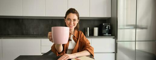 Stylish young woman, student studying from home, sitting in kitchen with homework, giving you cup of coffee, raising pink mug, smiling at camera photo