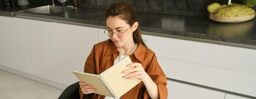 Close up portrait of young woman, student in kitchen, holding notebook, revising for exam at home, studying, reading her planner photo