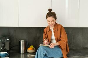 retrato de elegante joven mujer, se sienta a hogar en el cocina y hace notas, escribe abajo receta, cheques su lista con casa mandados, garabatear o dibujo en cuaderno foto