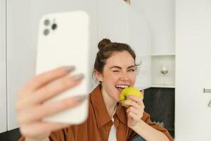 Portrait of happy, smiling young woman records herself, takes selfie while eating an apple in the kitchen, using smartphone app, makes photos with mobile phone