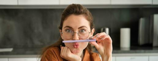 Close up portrait of funny, silly young woman, playing with pen, holding pencil on top of her lip and grimacing, sitting in kitchen photo