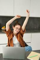 Vertical shot of smiling, lively young woman in glasses, working on remote, sitting at home with laptop, using computer to study online, stretching hands with pleased emotion photo