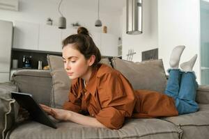 fin de semana y estilo de vida concepto. joven mujer acostado en sofá con digital tableta, desplazamiento social medios de comunicación, leyendo libro electronico o acecho televisión serie en aplicación foto