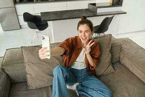 Portrait of cute smiling female model, takes selfie on smartphone app, posting photos on social media, using mobile phone camera, posing on sofa at home
