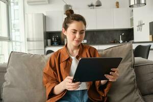 retrato de joven mujer sentado en sofá en vivo habitación, acecho películas en digital tableta, leyendo en dispositivo, disfrutando fin de semana a hogar foto