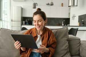 Portrait of young brunette woman reading on digital tablet, watching tv series on her application, sitting on couch in living room photo