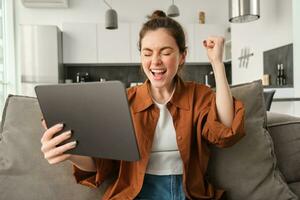 Portrait of excited young woman with digital tablet, sitting on couch, laughing and smiling, winning on video game, spending time at home photo