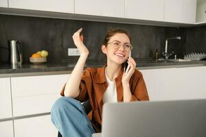 Image of beautiful modern woman working from home, studying in kitchen with laptop, talking on smartphone, calling someone on telephone photo