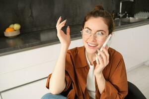 Excited young woman talking on mobile phone in front of laptop, sitting in kitchen with happy face expression, having a conversation photo