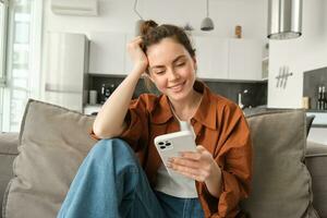 Portrait of happy young brunette woman sitting on sofa with smartphone, reading notification, messaging on mobile phone, resting on couch at home, smiling while looking at device screen photo