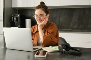 Portrait of young woman working from home, sets up workplace in kitchen, using laptop. Student doing homework on computer photo