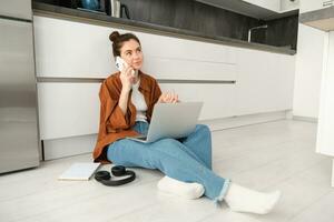 joven mujer por casualidad se sienta en piso con computadora portátil, hace un teléfono llamar, hablando en teléfono foto