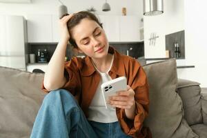 Portrait of beautiful young woman reading news on mobile phone, using smartphone app, looking at screen and smiling, sitting at home on sofa photo