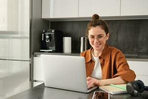 Portrait of young woman working from home, sets up workplace in kitchen, using laptop. Student doing homework on computer photo