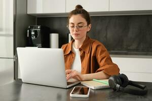 Work and life balance concept. Young woman in glasses, working from home, typing on laptop, student doing homework on computer, sitting in kitchen, freelancing photo