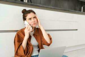 Portrait of woman calling her doctor on the phone, holding smartphone, frowning and touching her head, has painful headache or migraine, feeling unwell photo