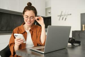 Portrait of young selfemployed woman, replying to customers on smartphone, using laptop to check orders, sitting at home photo