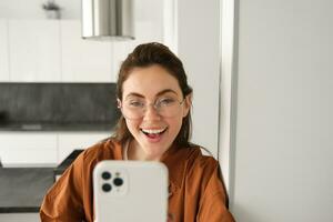 Close up portrait of happy girl takes selfie on smartphone, posing with mobile phone, looking excited and amazed, smiling at telephone camera, using social media app photo