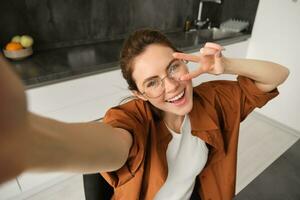 Selfie of happy young and carefree woman, taking photo on mobile phone with extended hand, posing and smiling, sitting in kitchen in glasses