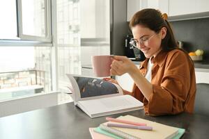 Portrait of woman reading at home, flipping pages of favourite book, relaxing in kitchen with cup of coffee, wearing glasses photo