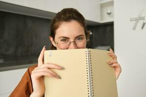 Close up portrait of cute brunette woman in glasses, sitting at home, smiling, hiding face behind yellow notebook photo