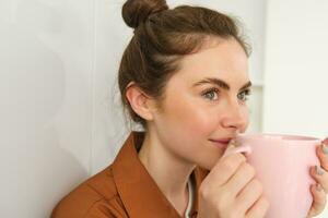 acogedor momentos contento joven mujer se sienta en cocina con taza de recién elaborada café, sonriente y mirando aparte. niña bebidas té a hogar foto