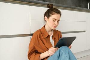 Portrait of pretty young woman at home, sitting on kitchen floor, looking at digital tablet, using gadget, reading on device photo