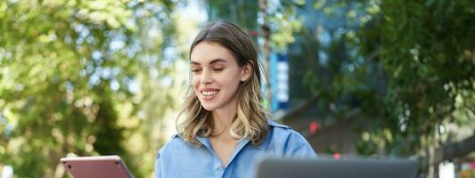 Vetical shot of young female student, reading on digital tablet, studying outdoors. Businesswoman sitting outside with laptop and gadgets, working photo