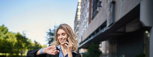 retrato de confidente mujer de negocios yendo en un reunión, en pie en calle, hablando en móvil teléfono y comprobación hora en digital reloj foto