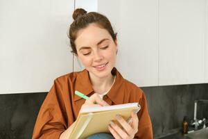 Portrait of cute young woman with notebook, sitting in the kitchen and writing her to do list, making tasks for week, puts notes in schedule or planner photo