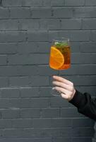 Close-up of woman's hand holding glass of cocktail with orange slice on black brick wall background photo