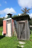 A rustic homemade shower with a barrel for heating water on the roof and a toilet are light wooden buildings in the garden of a rural house for the summer season in Russia. Vertical photo