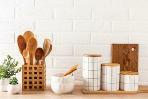 Set of stylish ceramic food storage jars, ceramic bowls, wooden spoons in a stand on a wooden countertop with potted indoor plants. photo