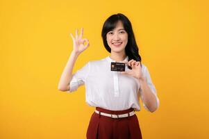 Portrait beautiful young asian woman enterpriser happy smile wearing white shirt and red plants showing okay hand gesture and holding credit card isolation on yellow background. photo