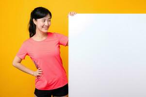 young asian sports fitness woman happy smile wearing pink sportswear standing behind the white blank banner or empty space advertisement board against yellow background. wellbeing lifestyle concept. photo