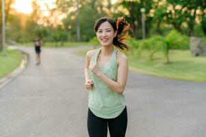 ajuste asiático joven mujer trotar en parque sonriente contento corriendo y disfrutando un sano al aire libre estilo de vida. hembra persona que practica jogging. aptitud corredor niña en público parque. sano estilo de vida y bienestar siendo concepto foto