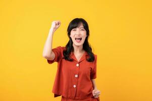 Portrait young beautiful asian woman happy smile dressed in orange clothes showing fist up hand gesture isolated on yellow studio background. Successful hooray celebrate young person concept. photo
