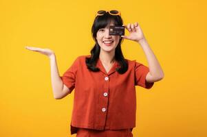 Portrait young beautiful asian woman happy smile dressed in orange clothes showing credit card isolated on yellow background. Pay and purchase shopping payment concept. photo