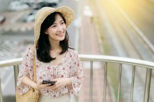 young asian woman traveler with weaving basket using mobile phone and standing on overpass with railway background. Journey trip lifestyle, world travel explorer or Asia summer tourism concept. photo