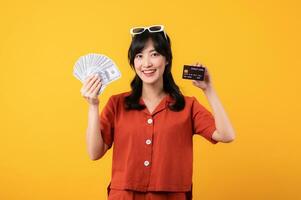 portrait of happy successful confident young asian woman happy smile wearing orange clothes holding cash money and credit card standing over yellow background. millionaire business, shopping concept photo