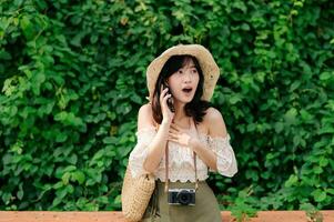 Portrait of asian young woman traveler with weaving hat, basket, mobile phone and camera on green public park background. Journey trip lifestyle, world travel explorer or Asia summer tourism concept. photo