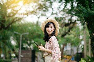 Portrait of asian young woman traveler with weaving hat and basket using mobile phone on green park nature background. Journey trip lifestyle, world travel explorer or Asia summer tourism concept. photo