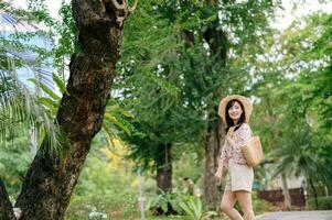 Portrait of asian young woman traveler with weaving hat and basket happy smile on green public park nature background. Journey trip lifestyle, world travel explorer or Asia summer tourism concept. photo