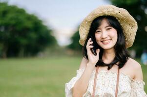 retrato de asiático joven mujer viajero con Costura sombrero, cesta, móvil teléfono y cámara en verde público parque antecedentes. viaje viaje estilo de vida, mundo viaje explorador o Asia verano turismo concepto. foto