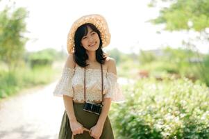 retrato de asiático joven mujer viajero con Costura sombrero y cesta y un cámara en verde público parque naturaleza antecedentes. viaje viaje estilo de vida, mundo viaje explorador o Asia verano turismo concepto. foto