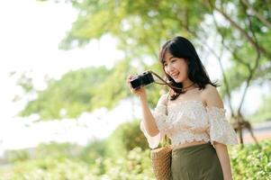 retrato de asiático joven mujer viajero con Costura sombrero y cesta y un cámara en verde público parque naturaleza antecedentes. viaje viaje estilo de vida, mundo viaje explorador o Asia verano turismo concepto. foto