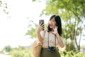 Portrait of asian young woman traveler with weaving hat, basket, mobile phone and camera on green public park background. Journey trip lifestyle, world travel explorer or Asia summer tourism concept. photo