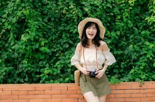 retrato de asiático joven mujer viajero con Costura sombrero y cesta y un cámara en verde público parque naturaleza antecedentes. viaje viaje estilo de vida, mundo viaje explorador o Asia verano turismo concepto. foto