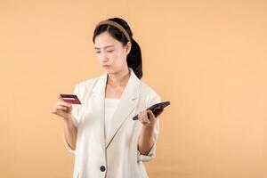 portrait of happy successful confident young asian business woman wearing white jacket holding smartphone and credit card standing over beige background. shopping concept. photo