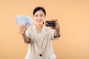 retrato de contento exitoso confidente joven asiático negocio mujer vistiendo blanco chaqueta participación efectivo dinero dolares y crédito tarjeta en pie terminado beige antecedentes. millonario negocio, compras concepto foto